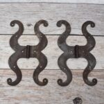 Pair of decorative black cast iron butterfly scroll hinges on a light wooden background.