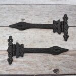 Pair of decorative black cast iron strap hinges with fleur-de-lis ends on a light wood plank background.