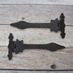 Pair of decorative black cast iron strap hinges with fleur-de-lis ends on a light wood plank background.