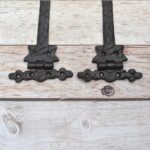 Pair of decorative black cast iron strap hinges with fleur-de-lis ends on a light wood plank background.