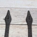 Pair of decorative black cast iron strap hinges with fleur-de-lis ends on a light wood plank background.