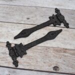 Pair of decorative black cast iron strap hinges with fleur-de-lis ends on a light wood plank background.
