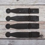 Set of four dark iron penny-end strap hinges on a light wood plank surface.