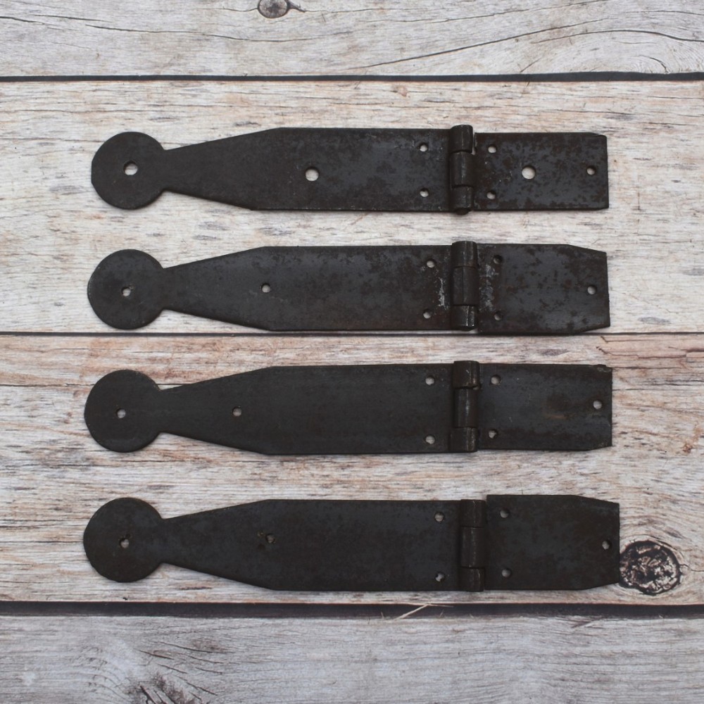 Set of four dark iron penny-end strap hinges on a light wood plank surface. Set of four dark iron penny-end strap hinges on a light wood plank surface.