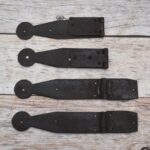 Set of four dark iron penny-end strap hinges on a light wood plank surface.