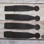 Set of four dark iron penny-end strap hinges on a light wood plank surface.