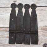 Set of four dark iron penny-end strap hinges on a light wood plank surface.